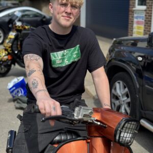 Model sitting on an orange GP wearing a black t-shirt with a green design featuring innocenti fuels.