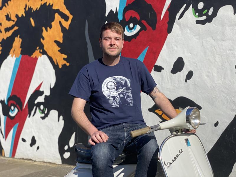 Man sitting on a white vintage vespa wearing a navy t-shirt with a scooter engine depicted as an x-ray inside a skull